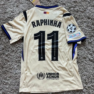Barcelona Raphinha 11 Nike fotbollströja - Snygg Barcelona fotbollströja med Raphinha 11 på ryggen. Tröjan är beige med svarta och blå detaljer, korta ärmar och tillverkad i Dri-Fit material. Officiella loggor från Nike, Spotify och UNHCR samt Champions League-märke på ärmen. Perfekt för dig som älskar Barca!