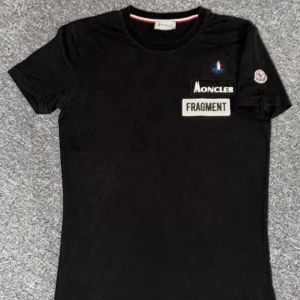 Svart Moncler Fragment t-shirt - Svart t-shirt från Moncler Fragment med broderade loggor och patches på bröstet och ärmen. 63cm längd och 44cm brädd se bild.Vid frågor eller behov av flera bilder hör av dig!