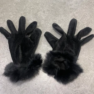 Svarta sammetshandskar med fuskpäls - Stilrena svarta handskar i mjuk sammet med fluffig svart fuskpäls runt handlederna. Perfekta för att ge din outfit en lyxig touch och hålla händerna varma under kyliga dagar. Snyggt och klassiskt snitt med fem fingrar.