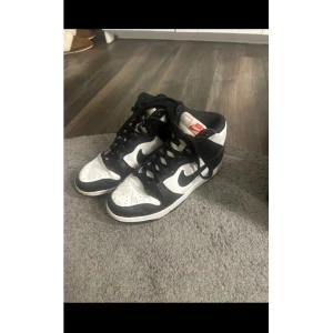 Nike Dunk High svartvita sneakers - Svartvita Nike Dunk High sneakers med klassisk design och röd Nike-logga på plösen. Skorna har snörning, perforerad tå och är tillverkade i skinn. Perfekta för dig som gillar streetstyle och vill ha en ikonisk look.
