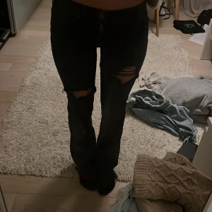 Svarta bootcut jeans med slitningar - Säljer ett par svarta jeans med bootcut-modell och snygga slitningar över knäna. Jeansen har klassisk femficksdesign och är tillverkade i ett stretchigt denimtyg som sitter skönt. Perfekta för dig som gillar en edgy och trendig look.