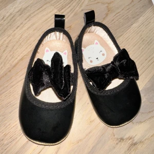 Svarta ballerinaskor med rosett H&M lacksko med rosett 🎁  - Söta svarta ballerinaskor från H&M med rund tå och mjuk platt sula. Skorna har en glansig finish / lacksko och en stor svart rosett framtill i sammet. Insidan har ett gulligt kattmotiv och en ljus skimrande bakgrund. Perfekta för att addera lite extra stil till outfiten.🤩✨