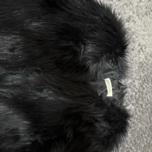 Svart pälsjacka med fluffig look - Säljer en svart pälsjacka med riktigt fluffig och mjuk känsla. Jackan har en klassisk krage och är helsvart med glansig finish. Perfekt för dig som vill sticka ut och hålla dig varm under kyliga dagar.