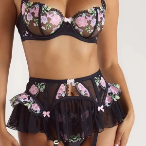 Svart blommig korsettopp från Lounge - Säljer ett svart set från Lounge med broderade blommor i rosa, lila och grönt. Toppen har spetsdetaljer, små rosetter och transparent meshmaterial. Justerbara axelband och dekorativa volanger ger en extra snygg look.