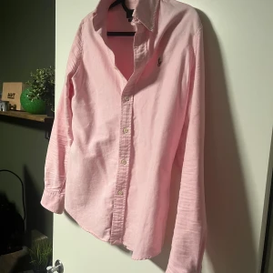 Rosa skjorta från Ralph Lauren - Snygg rosa skjorta från Ralph Lauren i custom fit med klassisk krage och knappar framtill. Skjortan har lång ärm och den ikoniska broderade loggan på bröstet. Tillverkad i mjuk bomull som känns skön mot huden. Perfekt för dig som gillar stilrena och fräscha plagg.