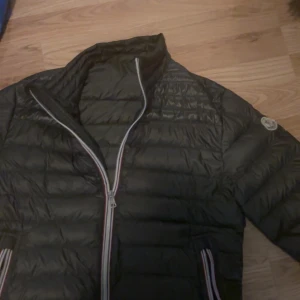  Moncler dunjacka herr 1 -  dunjacka från Moncler med klassisk quiltad design och Moncler-logga på ärmen. Jackan har dragkedja framtill och vid fickorna, samt diskreta ränder i rött, vitt och blått längs dragkedjorna. Perfekt för kyliga dagar och riktigt snygg streetstil. Pris diskuteras 
