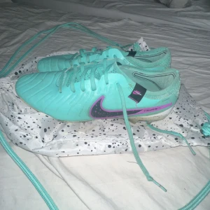 Nike Tiempo turkosa fotbollsskor - Säljer ett par Nike Tiempo fotbollsskor i en snygg turkos färg med lila och svarta detaljer. Skorna har snörning, rund tå och är tillverkade i syntetmaterial. Perfekta för dig som vill sticka ut på planen med stilrena och sportiga skor.