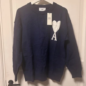 Mörkblå stickad tröja från Ami storlek M - Helt ny mörkblå stickad tröja från Ami Paris med vit hjärtapplikation och bokstaven A på bröstet. Tröjan har rund hals, ribbade muddar och lång ärm. Perfekt för dig som gillar stilrena detaljer och vill ha något som sticker ut.
