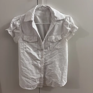 Vit kortärmad skjorta med puffärm - Snygg vit skjorta med korta puffärmar och klassisk krage. Skjortan har två bröstfickor med knappar och knäppning framtill. Perfekt för dig som gillar en clean och fräsch stil med lite extra detaljer på ärmarna.