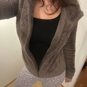 Brun teddyjacka från Sublevel - Mysig brun teddyjacka från Sublevel med huva och snörning med fluffiga bollar. Jackan har dragkedja framtill och långa ärmar. Perfekt för dig som gillar chill och comfy stil. Materialet är supermjukt och fluffigt, håller dig varm under kyliga dagar.