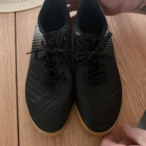Svarta inomhusfotbollsskor med mönster - Säljer ett par svarta inomhusfotbollsskor med coolt vitt mönster på hälen och klassisk gummisula. Skorna har snörning och är tillverkade i syntetmaterial, perfekta för snabba vändningar på planen.