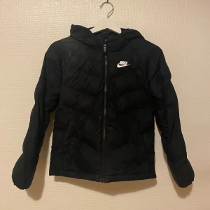 Svart Nike jacka - Svart pufferjacka från Nike med huva och vit logga på bröstet. Jackan har dragkedja framtill och är vadderad för extra värme. Perfekt för kyliga dagar och har en sportig look som funkar till det mesta.