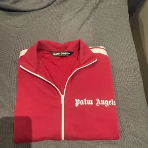 Röd track jacket från Palm Angels - Palm Angels röd track jacket med vit dragkedja och vita ränder på axlarna. Jackan har hög krage och logga tryckt på bröstet. Sportig och clean look, perfekt för streetwear-stil.