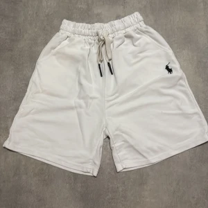 Vita shorts från Polo Ralph Lauren - Snygga vita shorts från Polo Ralph Lauren med klassisk broderad logga i svart på ena benet. De har elastisk midja med snörning och är tillverkade i mjuk bomull. Perfekta för en chill sommarlook.