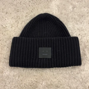Svart ribbstickad mössa Acne Studios - Svart ribbstickad mössa från Acne Studios med klassisk uppvikt kant och det ikoniska face-logotypen framtill. Perfekt till höst och vinter. Mycket fint skick bara lite nopprig på vissa ställen. Nypris 1700 kr.