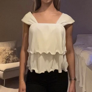 Vit plisserad blus med volang - Säljer en vit blus med plisserat tyg och dubbla volanger. Toppen har breda axelband och en snygg v-ringning både fram och bak. 