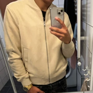 Beige bomberjacka från Zara - Snygg beige bomberjacka från Zara med stilren design och ribbade muddar. Jackan har dragkedja framtill, två sidofickor och en mjuk mockaliknande yta. Perfekt för dig som gillar minimalistisk och clean look.