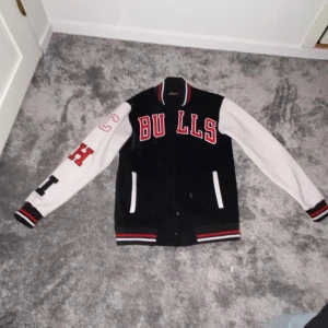 Svart och vit bomberjacka Bulls S - Snygg svart och vit bomberjacka från Sizelog i storlek S med stora röda BULLS-bokstäver på bröstet och broderade bokstäver på ena ärmen. Jackan har ribbade muddar med röd, vit och svart rand, knappar framtill och två fickor. Perfekt för dig som gillar sportig college-stil.