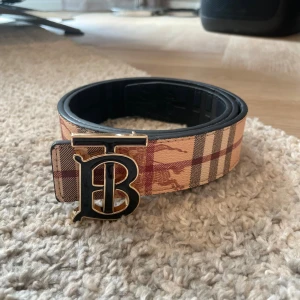 Burberry rutig skärp med guldspänne - Snyggt Burberry-bälte med klassiskt rutigt mönster i beige, rött och svart på ena sidan och svart med diskret mönster på andra. Stort guldfärgat TB-spänne framtill. Bältet är gjort i skinn och har en exklusiv känsla, perfekt för att lyfta vilken outfit som helst.