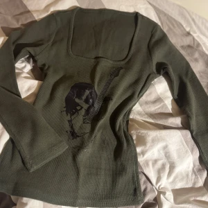 Olivgrön långärmad tröja med tryck - Olivgrön långärmad tröja med fyrkantig ringning och coolt svart tryck av en person med elgitarr på framsidan. Tröjan är ribbad och har en tight passform som framhäver siluetten. Perfekt för dig som gillar rockig stil.