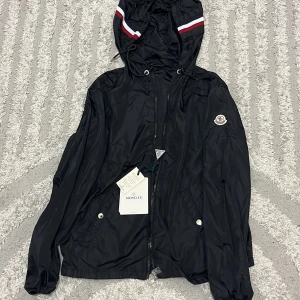 Svart vindjacka från Moncler med luva - Svart vindjacka från Moncler med luvaoch dragsko, dekorerad med röd och vit rand på huvan. Jackan har dragkedja framtill, två fickor med tryckknappar och Moncler-logga på ärmen. Tillverkad i glansig polyester, perfekt för en stilren look.