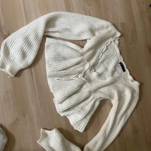 Vit stickad omlottopp med volang - Superfin vit stickad omlottopp med långa ärmar och volangkant längs urringningen. Toppen har knytband i midjan och är ribbstickad för en mysig känsla. Perfekt för dig som gillar romantiska detaljer och en feminin look.