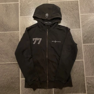 Svart hoodie från Sail Racing med dragkedja - Svart hoodie från Sail Racing med broderad logga och siffran 77 på bröstet. Tröjan har hel dragkedja, huva och är fodrad med mjuk fleece. Tillverkad i bomull och polyester, perfekt för kyliga dagar. Snygg och sportig stil med ribbade muddar.