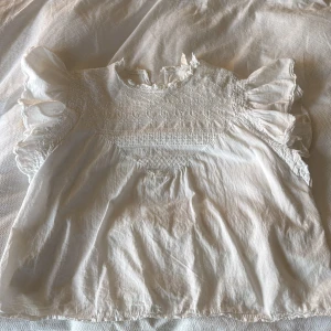 Vit blus med broderi från Auphoria - Supersöt vit blus från Auphoria med broderade detaljer och volangiga korta ärmar. Blusen har rund halsringning med liten volangkant, smock vid ärmslut och knäppning bak i nacken. 