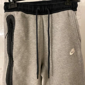Grå och svarta mjukisbyxor från Nike - Nike mjukisbyxor i grått med svarta detaljer och muddar. Byxorna har snörning i midjan, en stor dragkedja längs ena benet och Nike-logga på framsidan. Materialet är mjukt och bekvämt, perfekt för chill eller träning. Svarta partier nedtill ger en snygg kontrast.