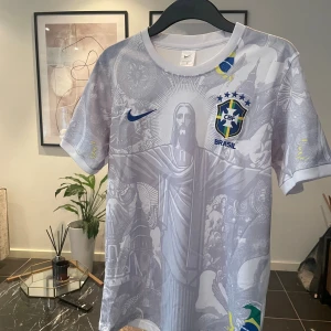 Vit Brasil Nike fotbollströja S - Säljer en vit Brasilien fotbollströja från Nike i storlek S. Tröjan har korta ärmar och ett detaljerat grafiskt mönster med Kristusstatyn och brasilianska symboler. Broderat CBF-emblem och Nike-logga i blått framtill. Materialet är lätt och andas, perfekt för match eller träning.