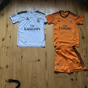 Real Madrid fotbollströjor Adidas - Två Real Madrid fotbollströjor från Adidas, en vit med svarta detaljer och Isco 23 på ryggen, samt en orange med svarta ränder och matchande shorts. Båda har klubbmärke och Fly Emirates-tryck. Klassisk sportdesign i lätt och ventilerande material. De vita shortsen är dolda separat till en tidigare kund.