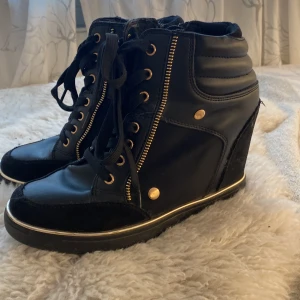 Svarta kilklackade sneakers med gulddetaljer - Snygga svarta sneakers med kilklack, guldfärgade dragkedjor och nitar. Skorna har snörning framtill, rund tå och detaljer i både skinn och mocka. Perfekta för dig som vill ha en sportig look med lite extra höjd och stil.