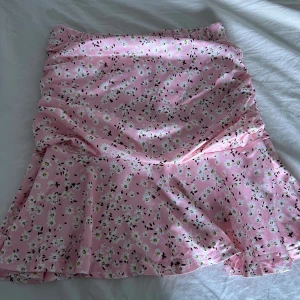 Rosa blommig kortkjol med volang - Supersöt rosa kortkjol med små vita blommor och volangkant. Kjolen har en lätt utsvängd form och är perfekt för vår och sommar. Materialet känns mjukt och följsamt mot huden.