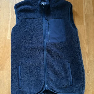 Mörkblå fleeceväst med dragkedja - Mysig mörkblå fleeceväst med hög krage och hel dragkedja framtill. Västen har två sidofickor och en bröstficka med dragkedja, perfekt för att hålla småsaker säkra. Materialet är mjukt och fluffigt, vilket gör den skön att bära under kyliga dagar. Storlek 160 nästan lika stor som xs. Kan piffa till din outfit rejält.