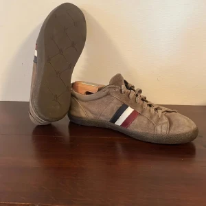 Bruna skor från Moncler i mocka - Snygga bruna skor från Moncler i mjuk mocka med klassiska ränder i rött, vitt och blått på sidan. Skorna har snörning, rund tå och låg profil. Perfekta för dig som vill ha en stilren och sportig look med exklusiv känsla. Pris inte spikat våga buda! Passar större i strlk. Dustbag ingår!