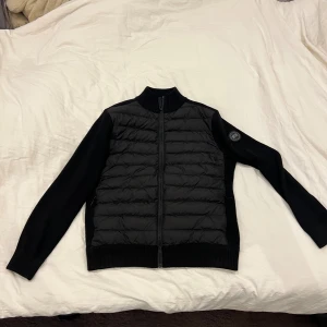 Svart cardigan från Canada Goose - Snygg svart jacka från Canada Goose med quiltad front och stickade ärmar. Jackan har dragkedja hela vägen, ribbad krage och diskret logga på ärmen. Perfekt för dig som vill ha en stilren och modern look med premiumkänsla. Priset kan diskuteras vid snabb affär 