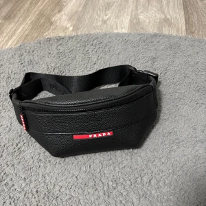 Svart axelväska i skinn - Snygg svart axelväska i grymt mjukt och strukturerat skinn. Väskan har ett brett justerbart band med logga och en dragkedja med röd detalj. Perfekt storlek för det viktigaste och stilren design med tydlig branding.