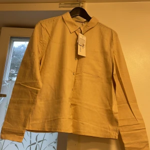 Beige skjorta från Anna Field - Stilren beige skjorta från Anna Field i 100% bomull. Skjortan har klassisk krage, knäppning framtill och en bröstficka. Långärmad och med rak passform, perfekt för en clean och enkel look.