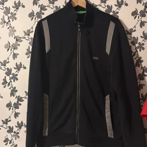 Svart kofta från Hugo Boss - Svart kofta från Hugo Boss med gråa ränder längs axlar och sidor. Kofta har hel dragkedja framtill, hög krage och diskret Boss-logga på bröstet. Sportig och stilren design, perfekt för en clean streetwear-look.