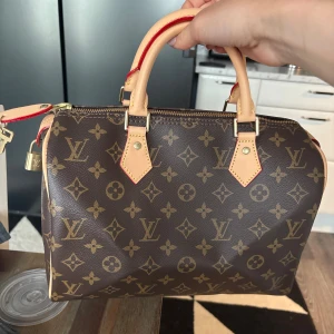 Louis Vuitton Speedy Monogram väska - Säljer en klassisk Louis Vuitton Speedy handväska i brunt monogrammönster med beigea handtag och detaljer. Väskan har guldfärgade metalldetaljer, dragkedja och ett rymligt huvudfack med innerficka. Perfekt för dig som gillar lyxiga accessoarer.