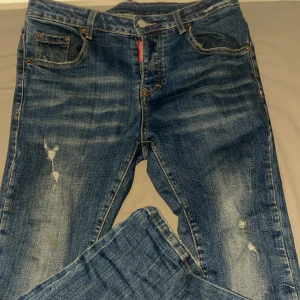 Blå jeans från Dsquared2 med slitningar - Snygga blå jeans från Dsquared2 med coola slitningar och en klassisk femficksdesign. Jeansen har en orange läderpatch bak i midjan och tydliga kontrastsömmar. Passar dig som gillar streetstyle och vill sticka ut med detaljer.