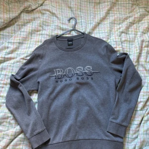 Grå slim fit tröja från Hugo Boss - Säljer en grå långärmad tröja från Hugo Boss med stilren BOSS-logga framtill. Tröjan har rund halsringning, ribbade muddar och är i slim fit-modell. Perfekt för dig som gillar en clean och klassisk look.