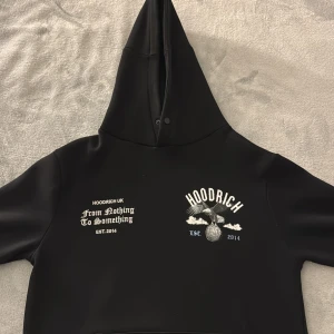 Svart hoodie från Hoodrich med tryck - Svart hoodie från Hoodrich i storlek S med tryck både fram och bak. Framsidan har text och logga, medan baksidan har ett stort tryck med örn, moln och mönster. Hoodien har huva.