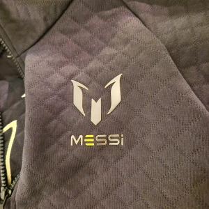 Svart Messi hoodie från Adidas - Säljer en svart quiltad hoodie från Adidas med Messi-logga på bröstet och ryggen. Tröjan har huva, half zip-dragkedja och praktisk magficka. Perfekt för dig som gillar fotboll och vill ha en snygg och sportig look.den som köper behöver betala för frakt det kommer kosta mer om den ska utt från eu
