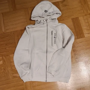 Vit hoodie från Sail Racing med dragkedja - Vit hoodie från Sail Racing med hel dragkedja framtill, huva med snörning och en bröstficka med dragkedja. Snygg logga på bröstet och på huvan. Mjuk insida och ribbade muddar vid ärmslut och nederkant.