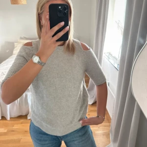 Ljusgrå stickad tröja med cut out - Säljer en ljusgrå stickad tröja med trendiga cut out-detaljer på axlarna. Tröjan har halvlånga ärmar och en klassisk rund halsringning. Perfekt för dig som vill ha en enkel men ändå unik look.