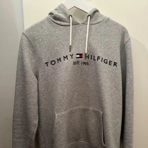 Grå hoodie från Tommy Hilfiger M - Snygg grå hoodie från Tommy Hilfiger i ekologisk bomull. Klassisk modell med huva och dragsnören, stor magficka och broderad logga på bröstet samt liten flagga på ärmen. Perfekt för chill dagar och enkel att matcha med jeans eller joggers. Lite. Fläck går bort i tvätten