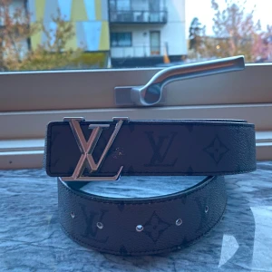 Louis Vuitton svart skärp med monogram - Svart Louis Vuitton-bälte i skinn med klassiskt monogrammönster och stor silvrig LV-logga som spänne. Bältet har en stilren och lyxig känsla, perfekt för att lyfta vilken outfit som helst. Tillverkat i Frankrike och har flera justerbara hål.