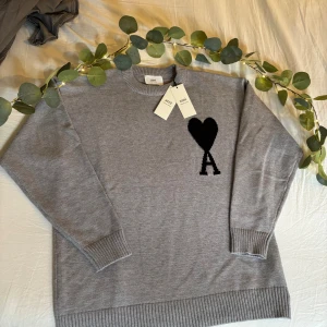 Grå stickad tröja från Ami Paris - Grå stickad tröja från Ami Paris med svart hjärta och A-logga på bröstet. Klassisk rund halsringning och ribbade muddar vid ärmslut och nederkant. Perfekt för dig som gillar stilrena och trendiga plagg med en twist.
