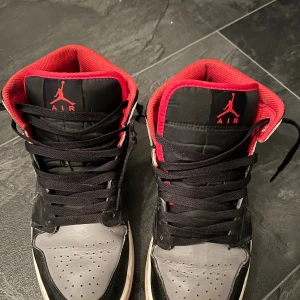 Nike Air Jordan 1 svart/röd/grå US8 - Säljer ett par Nike Air Jordan 1 i svart, grått och rött. Klassisk high-top siluett med svart snörning, röd insida och röd yttersula. Skorna har ikoniska Jordan Wings-loggan på sidan och är tillverkade i skinn och syntet. Perfekta för dig som gillar streetwear och sneakerskultur.